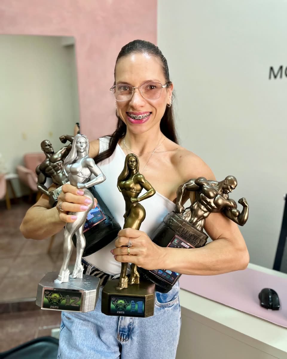 Monalysa Rodrigues de óculos, sorrindo no consultório e segurando quatro troféus de fisiculturismo